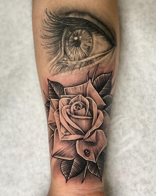 A black and grey realism tattoo on the forearm featuring a detailed eye above a large rose with intricate shading — by imindisoul, los angeles