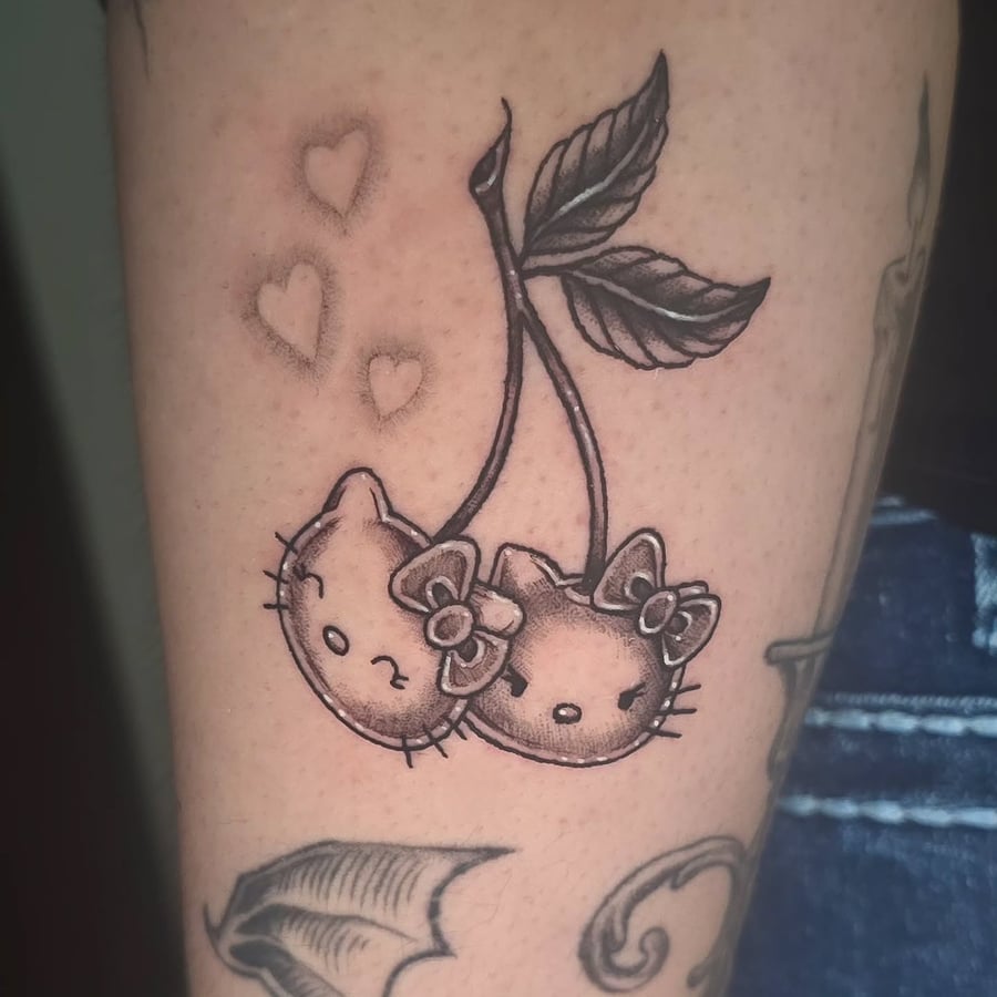 A whimsical black and grey tattoo of cherries with cat faces on the arm, featuring delicate heart accents — by Miguel ANGELO, los angeles