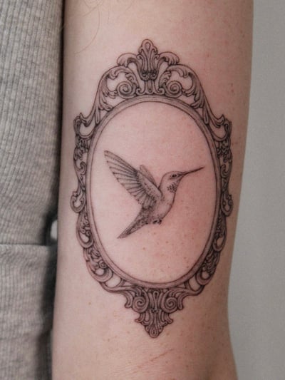A delicate black and grey fine line tattoo of a hummingbird inside an ornate frame on the arm — by Melanie Benoit, los angeles