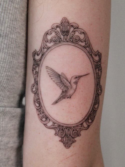 A delicate black and grey fine line tattoo of a hummingbird inside an ornate frame on the arm — by Melanie Benoit, los angeles