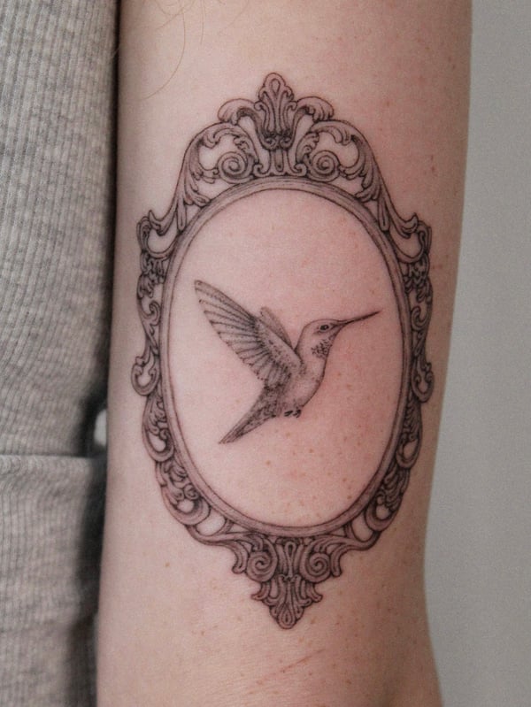 A delicate black and grey fine line tattoo of a hummingbird inside an ornate frame on the arm — by Melanie Benoit, los angeles