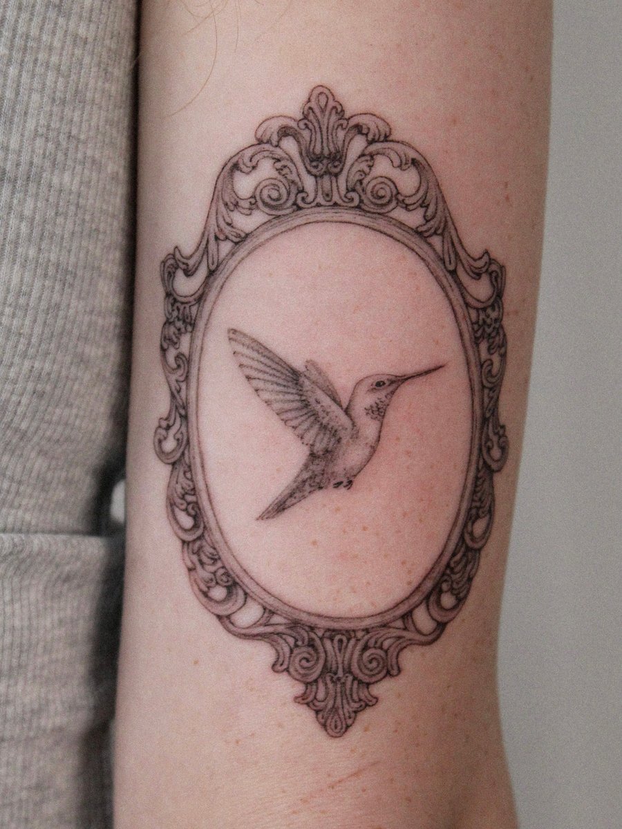 A delicate black and grey fine line tattoo of a hummingbird inside an ornate frame on the arm — by Melanie Benoit, los angeles