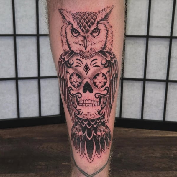 A bold black and grey tattoo of an owl with a sugar skull design on the calf, featuring intricate illustrative details — by Miguel ANGELO, temecula, ca