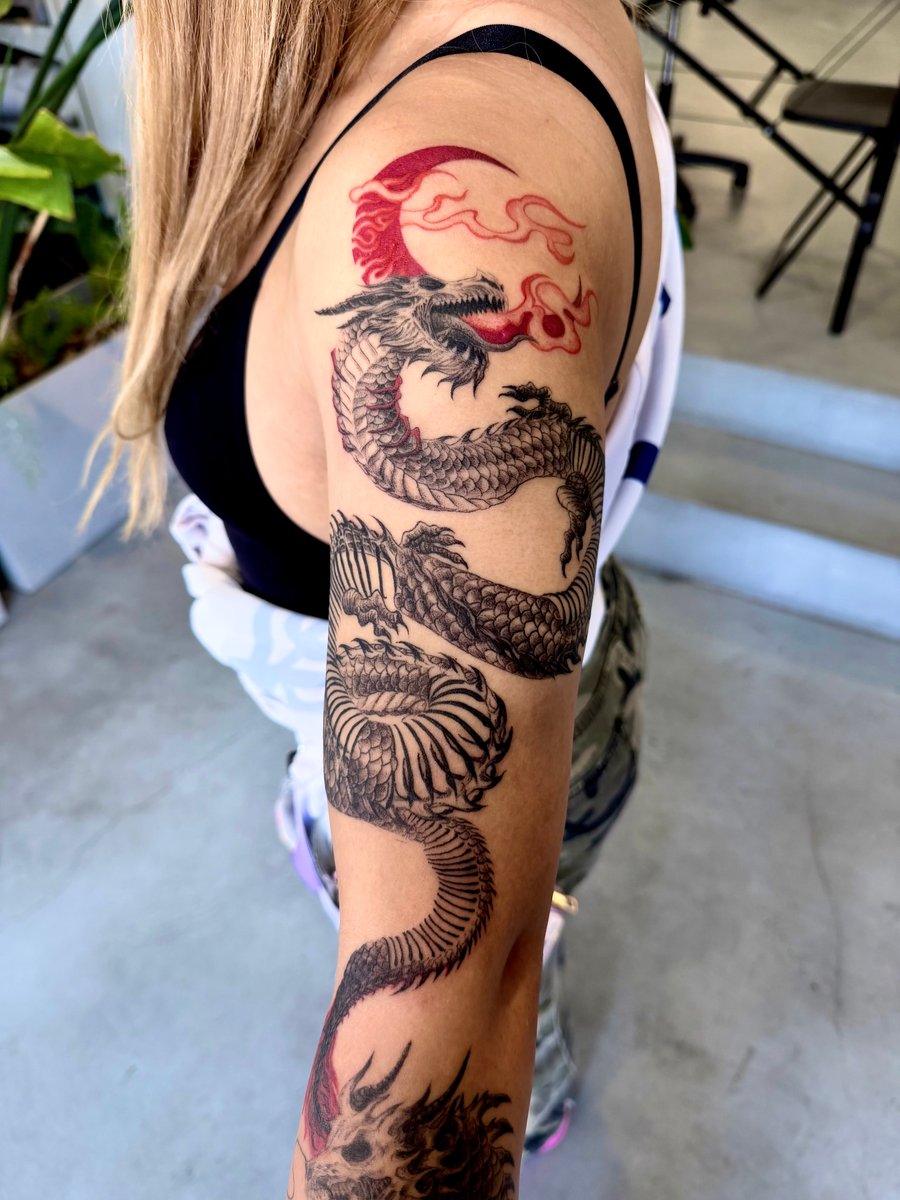 A Japanese-style dragon tattoo on the arm, featuring intricate black and grey scales with a red flame accent near the shoulder — by zitronxus, los angeles
