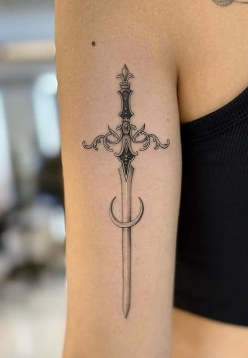A fine line black and grey tattoo of an ornate sword on the upper arm, featuring intricate detailing and elegant design — by otaybeatdown, los angeles