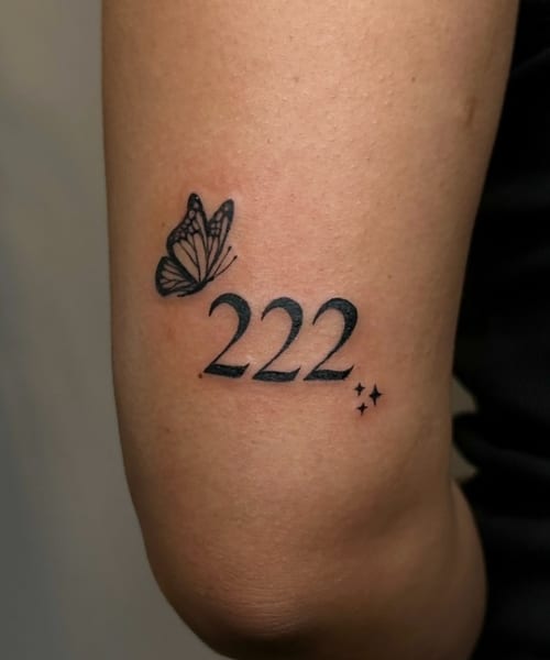 A minimalist blackwork tattoo on the upper arm featuring a butterfly and the number '222' with small stars — by NJ TATTOOZ, los angeles