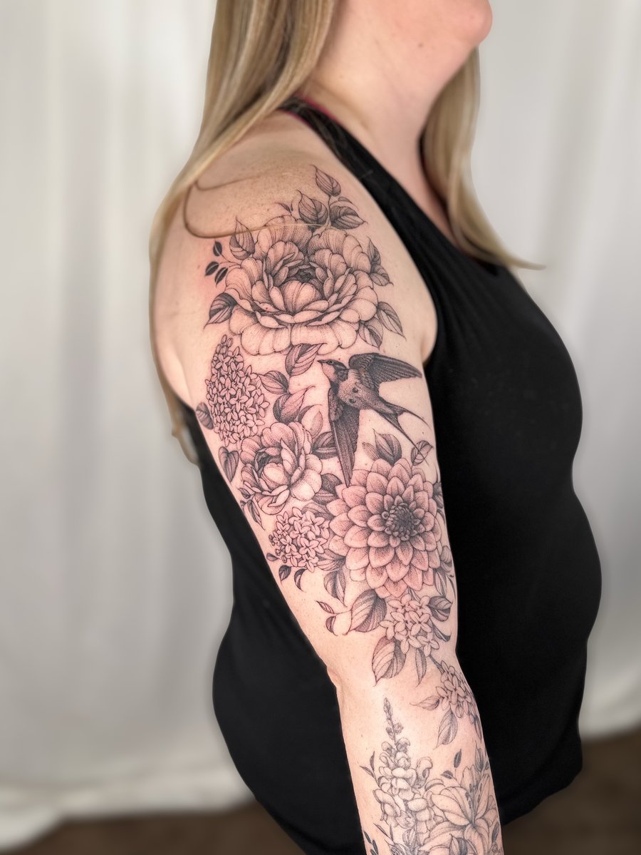 A detailed black and grey tattoo of a bird and floral arrangement on the upper arm, featuring intricate flowers and leaves — by Melanie Benoit, los angeles