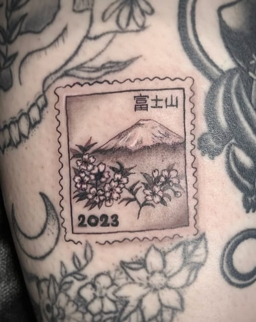 A black and grey illustrative tattoo of a postage stamp featuring a mountain and flowers, dated 2023, on the leg — by Shelby Bianco, los angeles