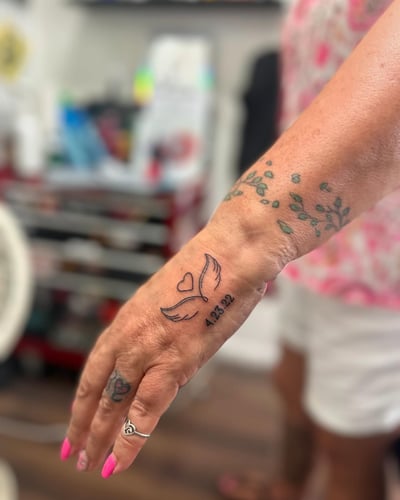 A delicate blackwork tattoo of a heart with wings and a date on the hand — by Maddie Ramirez, los angeles