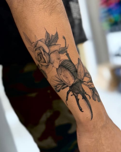 A black and grey tattoo on the forearm depicting a realistic beetle and a rose, with detailed linework and shading — by otaybeatdown, los angeles