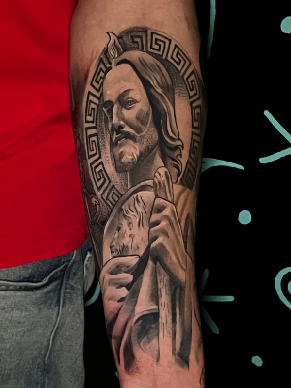 A realistic black and grey portrait of a religious figure on the forearm, featuring intricate shading and a geometric halo — by Josue Bravo, los angeles