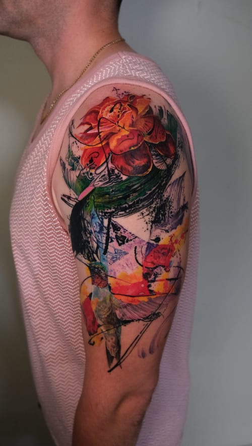 A vibrant watercolor tattoo on the upper arm featuring abstract floral elements with splashes of color and dynamic brushstroke effects — by Nik Lucas, los angeles
