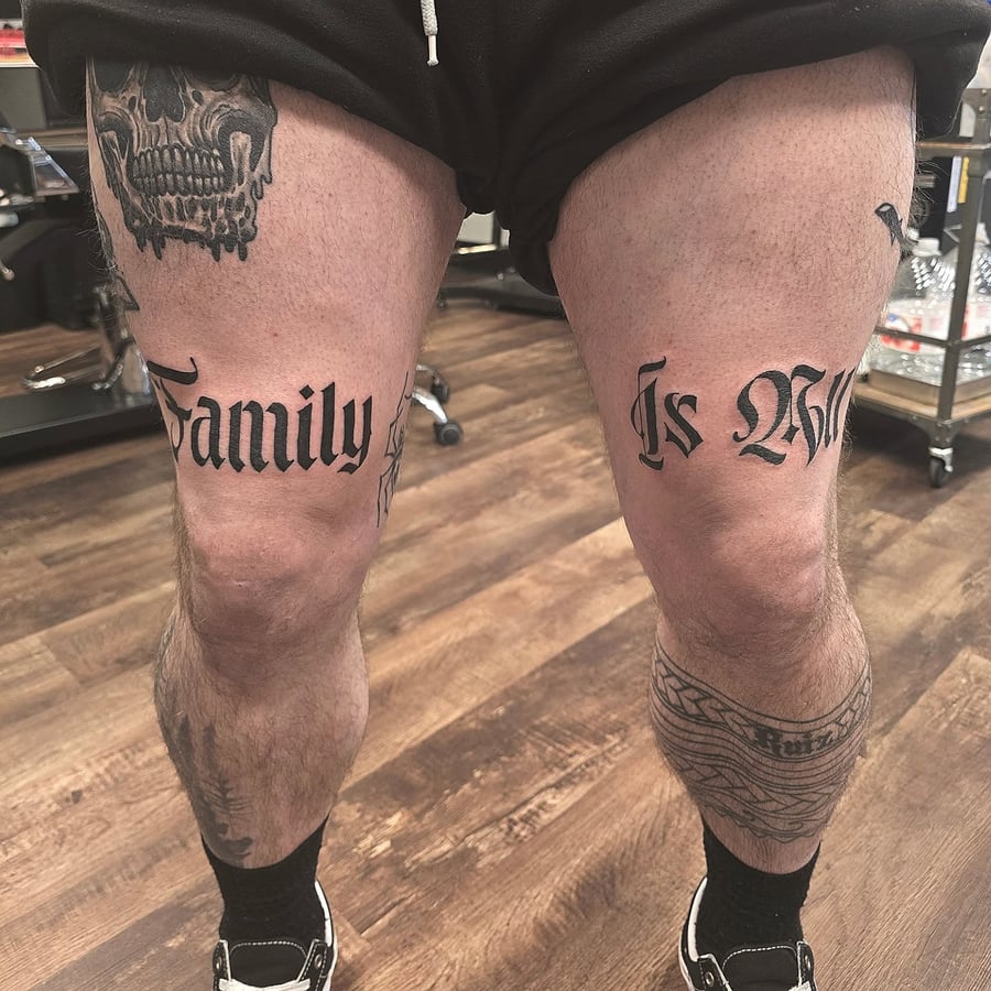 A bold black and grey script lettering tattoo on the thighs reading 'Family Is All' with a strong emphasis on family values — by Miguel ANGELO, los angeles