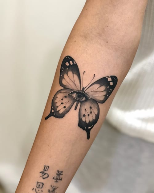 A surreal black and grey tattoo of a butterfly with an eye on the forearm, combining realistic and imaginative elements — by otaybeatdown, los angeles