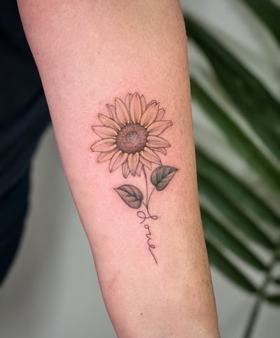 A delicate fine line sunflower tattoo on the forearm with a script word forming the stem, featuring soft color shading — by Melanie Benoit, los angeles