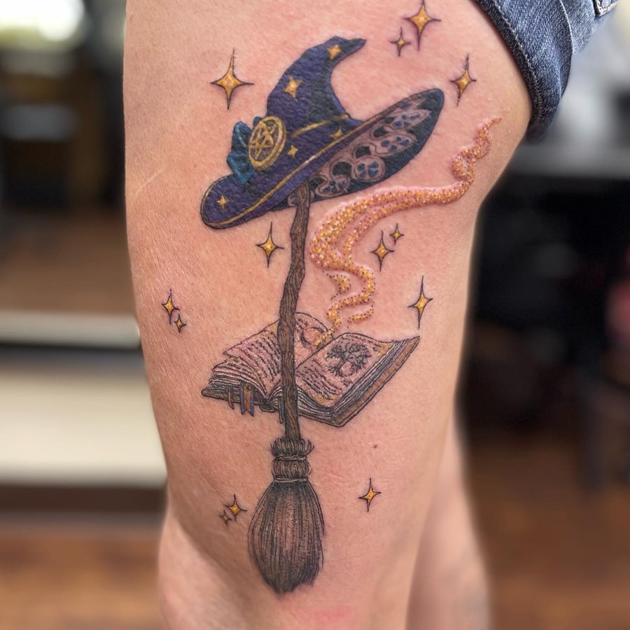 A colorful tattoo of a witch's hat, broom, and book on the thigh, featuring whimsical stars and magical elements — by Sandra Price, los angeles