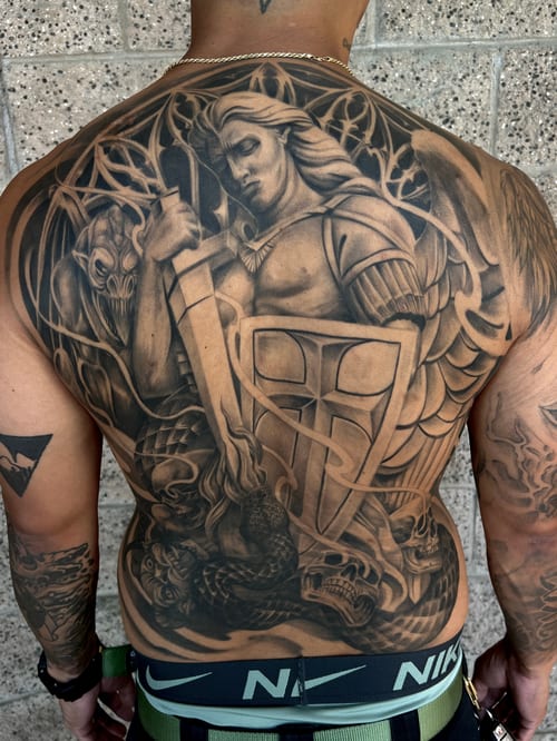 A detailed black and grey realism tattoo of Archangel Michael battling a demon, covering the full back with intricate shading and mythological elements — by imindisoul, los angeles