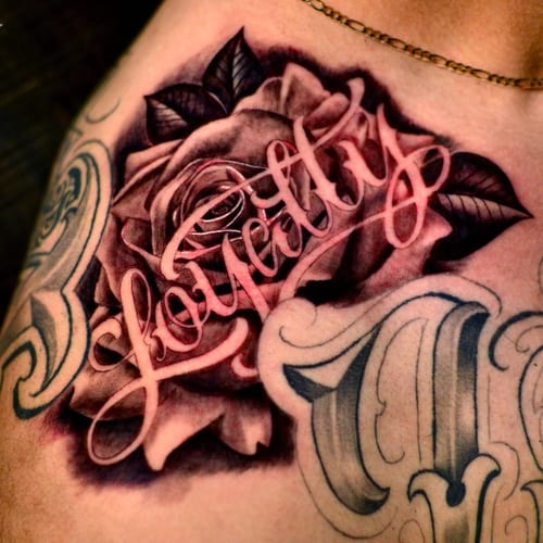 A bold black and grey tattoo of a rose with ornate script lettering spelling 'loyalty' on the shoulder — by Johnny Ayala, los angeles
