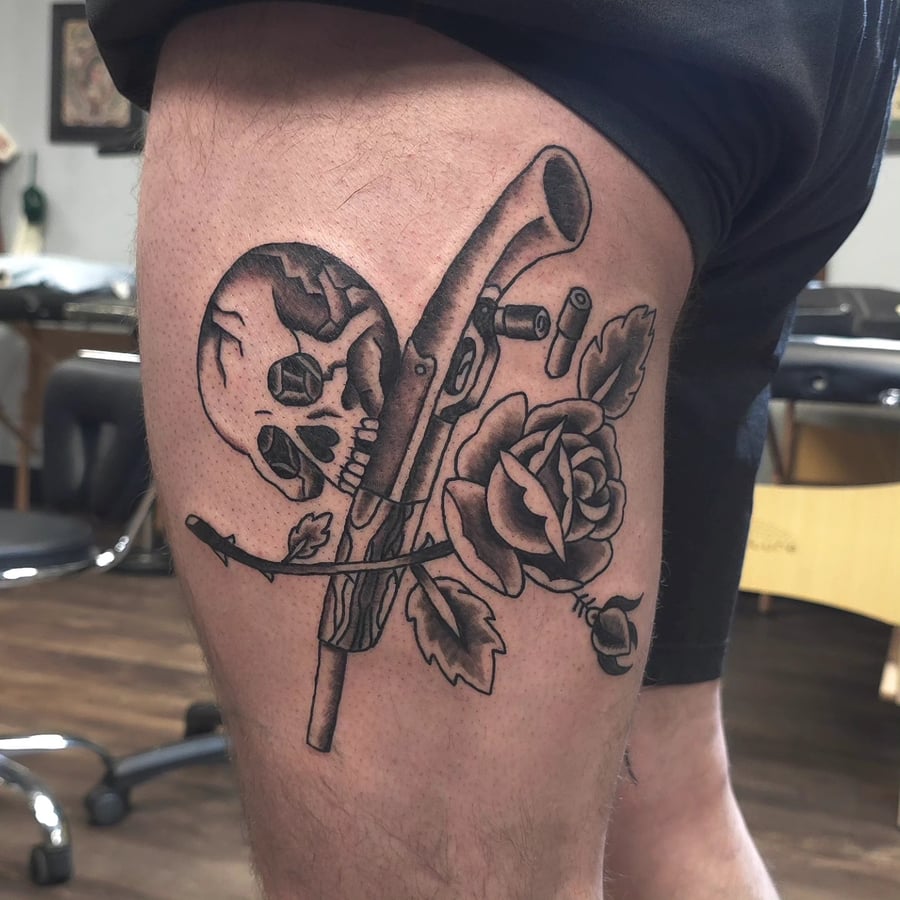 A bold black and grey tattoo on the thigh featuring a skull, rose, and gun in traditional style — by Miguel ANGELO, los angeles