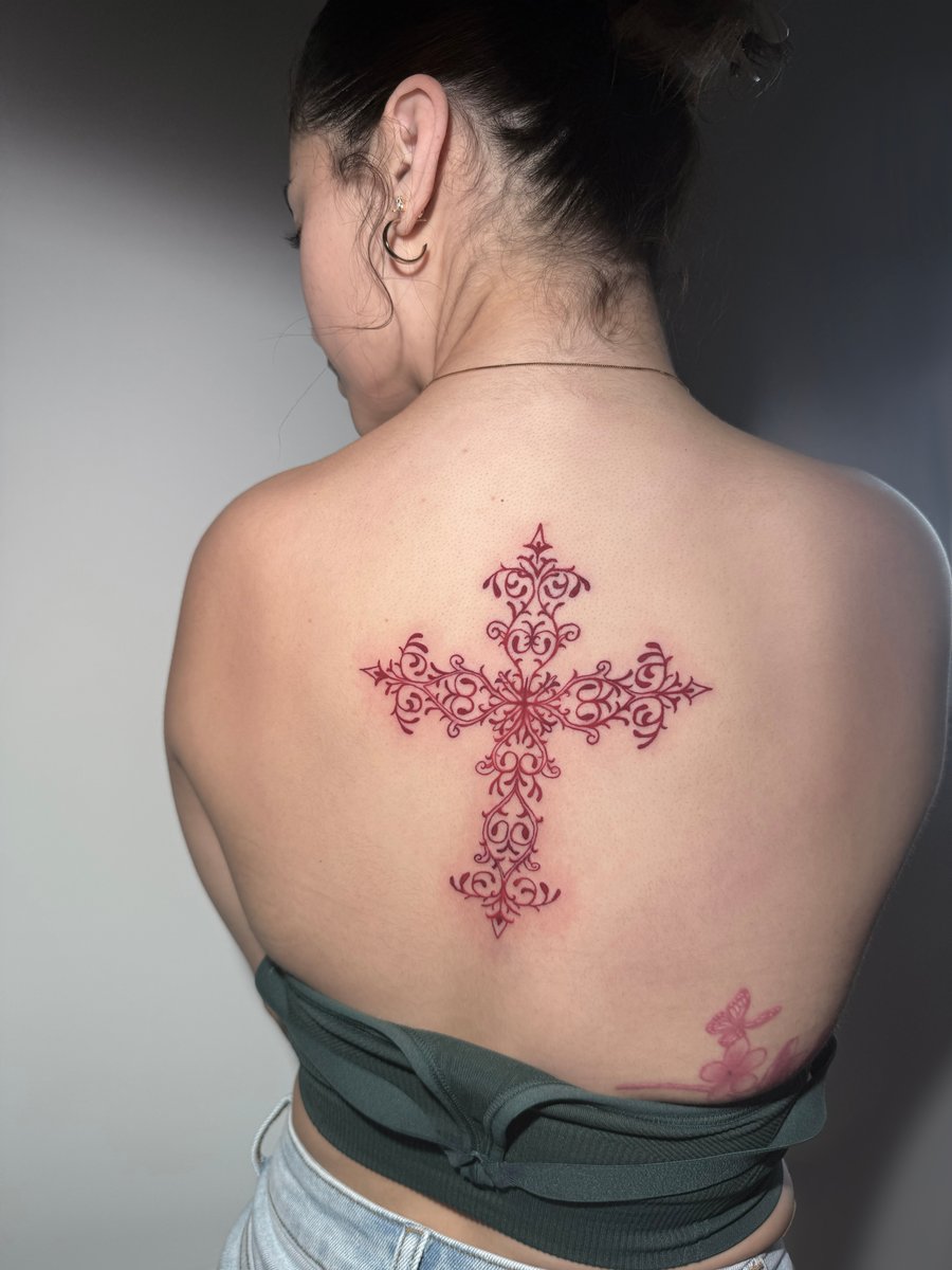An ornamental cross tattoo in red ink on the upper back, featuring intricate patterns and symmetrical design — by Shelby Bianco, los angeles