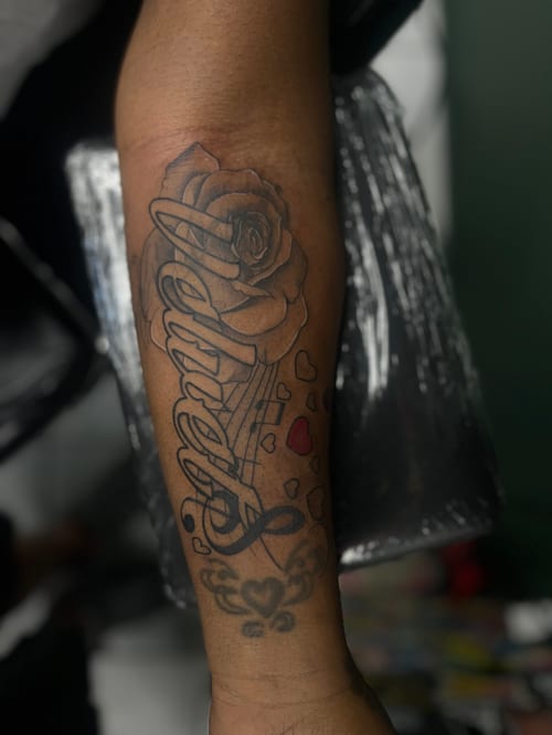 A forearm tattoo featuring a rose and the word 'Angel' in bold lettering, with small red hearts, combining blackwork and lettering styles — by Mr._Folks, los angeles