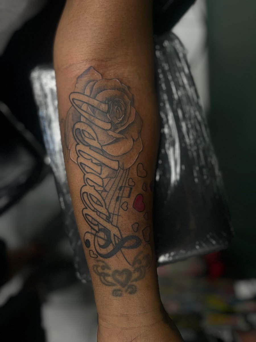 A forearm tattoo featuring a rose and the word 'Angel' in bold lettering, with small red hearts, combining blackwork and lettering styles — by Mr._Folks, los angeles