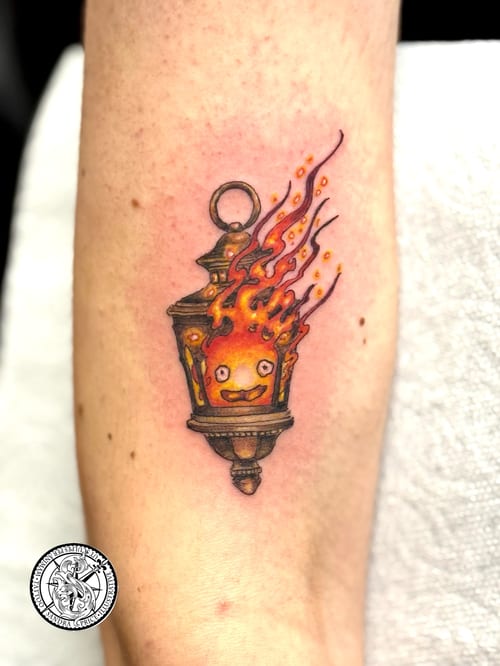 A whimsical and colorful tattoo of a flame with a face inside a lantern on the forearm — by Sandra Price, los angeles