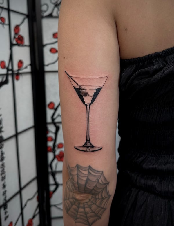 A black and grey realism tattoo of a martini glass on the upper arm, featuring detailed reflections and shading — by emily, rahway