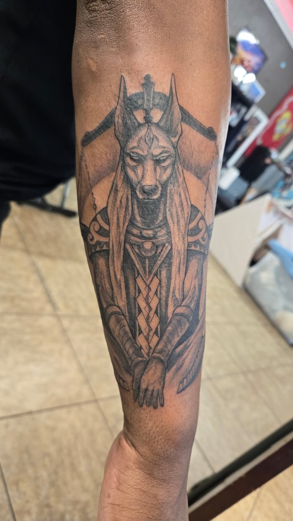 A detailed black and grey realism tattoo of Anubis on the forearm, featuring intricate shading and mythological elements — by Dretty, long beach, ca