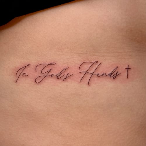 A delicate fine line script tattoo on the rib reading 'In God's Hands' with a small cross, done in black ink — by NJ TATTOOZ, los angeles