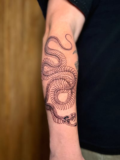 A detailed black and grey snake skeleton tattoo on the forearm, featuring intricate linework and a graphic style — by Dylan Aiello, los angeles