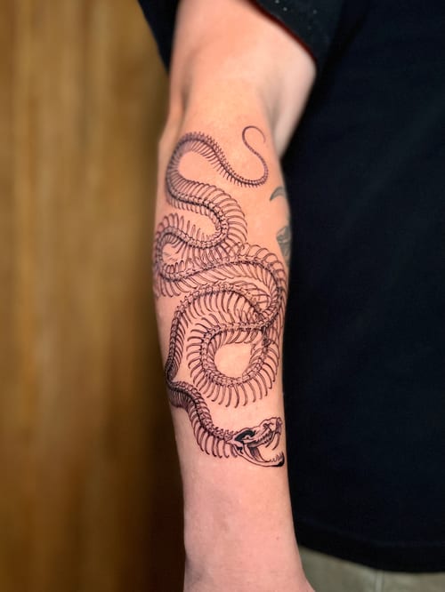 A detailed black and grey snake skeleton tattoo on the forearm, featuring intricate linework and a graphic style — by Dylan Aiello, los angeles