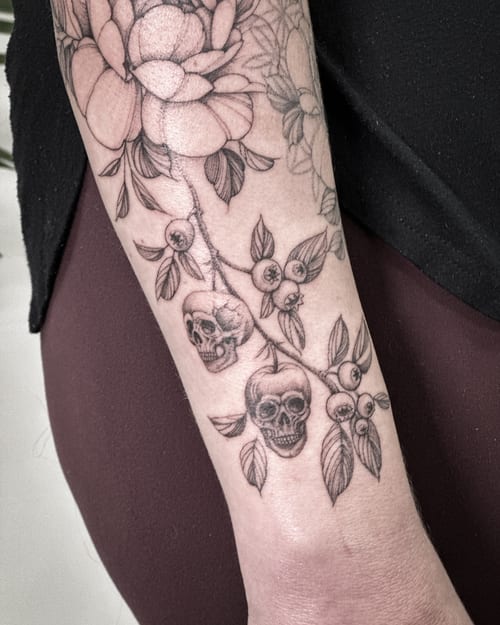 A black and grey tattoo of skulls intertwined with flowers on the forearm, featuring detailed blackwork and botanical elements — by Melanie Benoit, los angeles