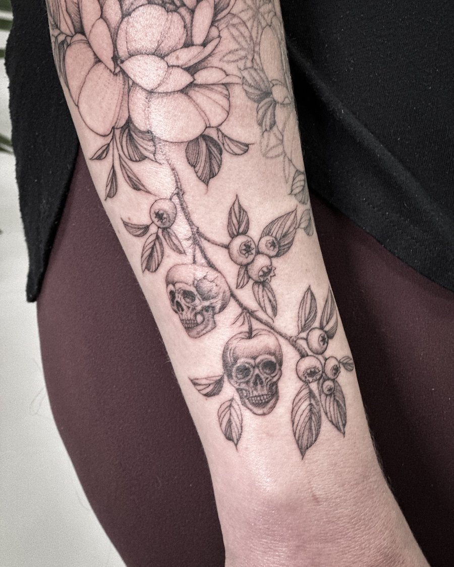 A black and grey tattoo of skulls intertwined with flowers on the forearm, featuring detailed blackwork and botanical elements — by Melanie Benoit, los angeles
