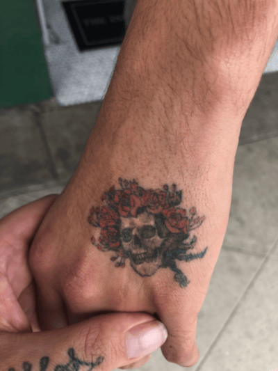 A small neo-traditional tattoo of a skull surrounded by red roses on the hand, featuring bold outlines and vibrant colors — by tyler_whitlock, los angeles