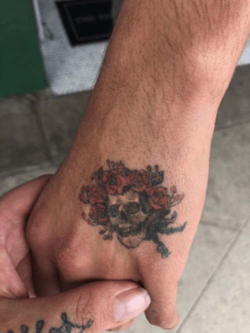 A small neo-traditional tattoo of a skull surrounded by red roses on the hand, featuring bold outlines and vibrant colors — by tyler_whitlock, los angeles