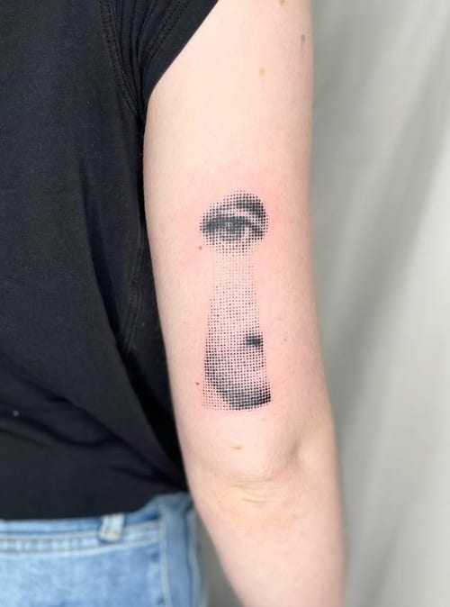 A black and grey dotwork tattoo of a face partially visible through a keyhole shape on the upper arm, creating a surreal effect — by Level tattoo, los angeles