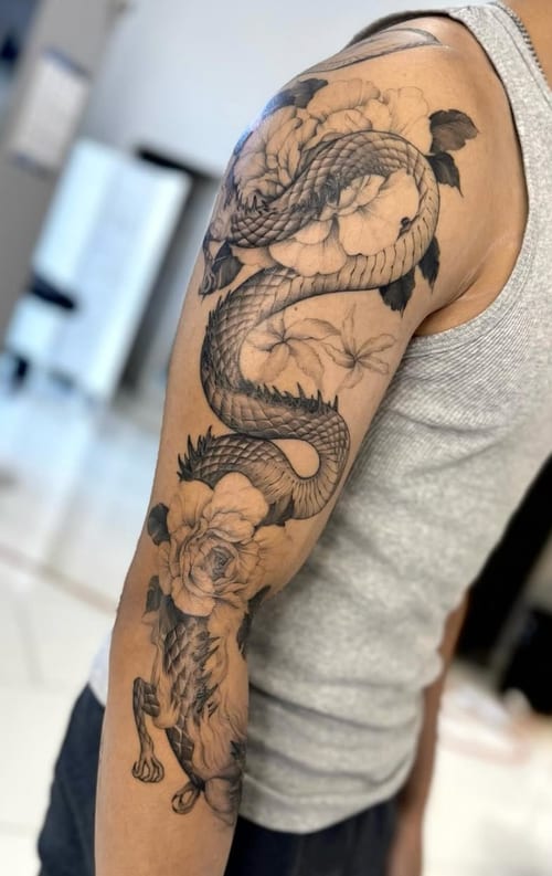 A black and grey Japanese-style dragon intertwined with flowers on the arm, featuring intricate details and smooth shading — by otaybeatdown, los angeles