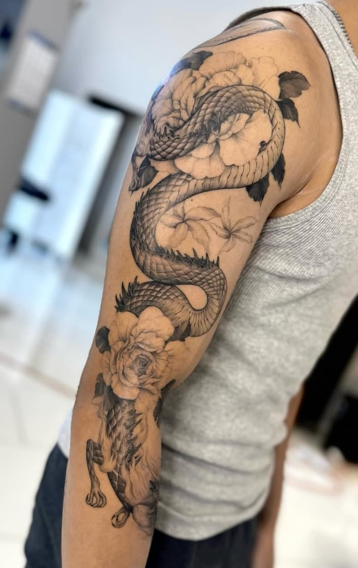 A black and grey Japanese-style dragon intertwined with flowers on the arm, featuring intricate details and smooth shading — by otaybeatdown, los angeles