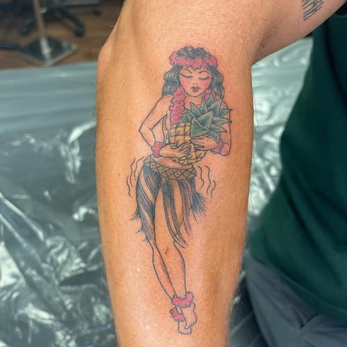 A colorful traditional style tattoo of a hula dancer holding a pineapple on the forearm — by Sandra Price, los angeles