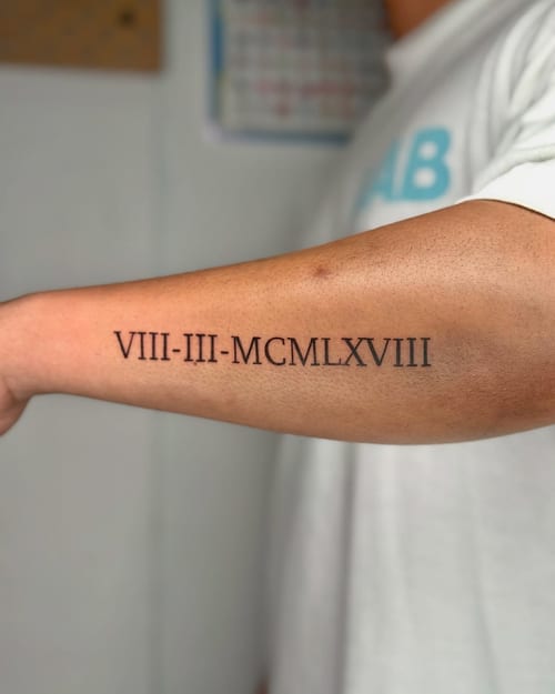 A minimalist black ink roman numeral tattoo on the forearm, featuring a clean and simple design — by zealous, los angeles