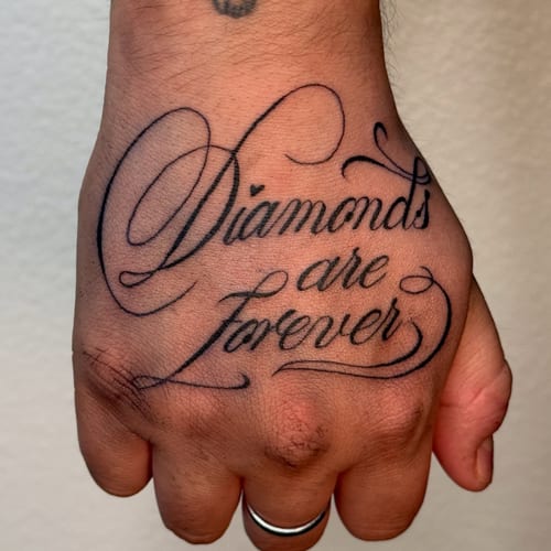 A bold blackwork script lettering tattoo on the hand reading 'Diamonds are Forever' with clean lines and elegant curves — by NJ TATTOOZ, los angeles