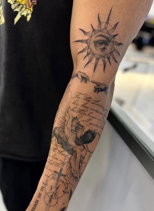A black and grey tattoo on the forearm featuring a realistic eye within a sun, an angel, and script lettering. The design is detailed and spans a significant portion of the forearm — by otaybeatdow…