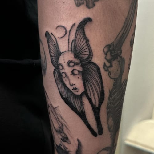 A surreal black and grey moth with a human face tattoo on the forearm — by Maddie Ramirez, los angeles