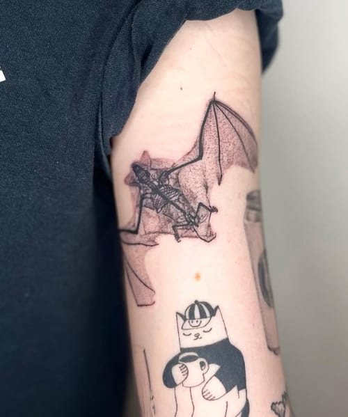 A black and grey linework tattoo of a bat with a skeleton on the upper arm, featuring detailed outlines and shading — by Level tattoo, los angeles