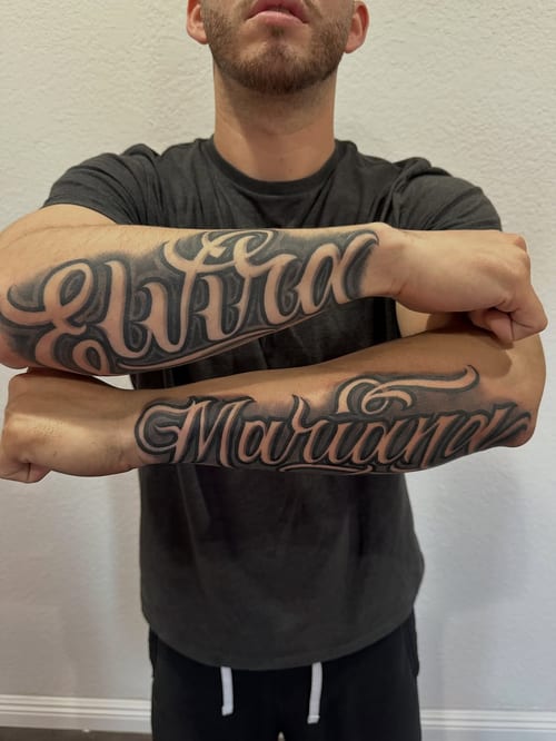 Bold black and grey script lettering tattoo on the forearms, featuring names in an ornate style — by Johnny Ayala, los angeles