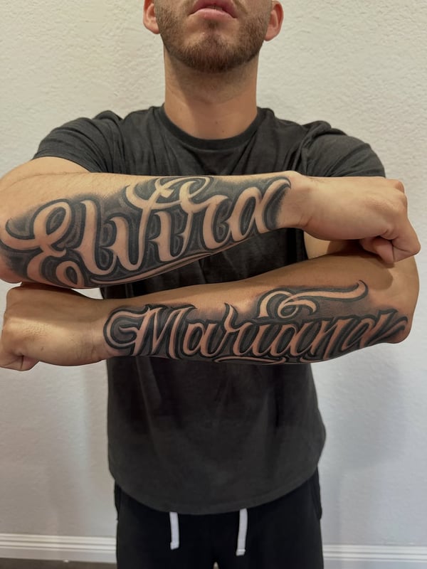 Bold black and grey script lettering tattoo on the forearms, featuring names in an ornate style — by Johnny Ayala, fontana, ca
