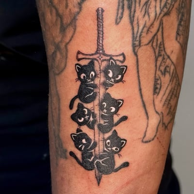 A whimsical black and grey tattoo of black cats climbing a sword on the forearm, done in an illustrative style — by NJ TATTOOZ, los angeles