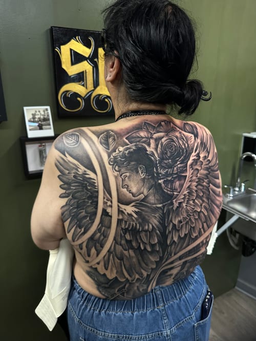 A detailed black and grey tattoo of an angel with wings and a rose on the full back, featuring realistic shading and intricate details — by Humilde tattoos, los angeles