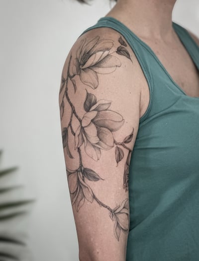 A delicate black and grey magnolia flower tattoo on the upper arm with detailed floral design — by Melanie Benoit, los angeles